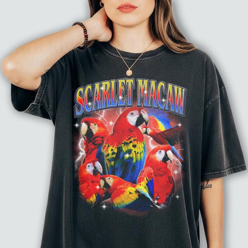 May include: Black t-shirt featuring a vibrant graphic of scarlet macaws with red, yellow, blue, and green plumage. The text "SCARLET MACAW" is displayed above the birds in a colorful, retro font, with lightning bolts in the background.