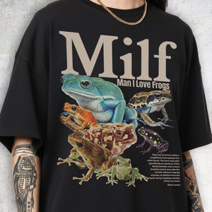 May include: Black t-shirt featuring a colorful graphic of several frogs. The text "Milf Man I Love Frogs" is printed above the frogs. A small paragraph about frogs is printed below the frogs.