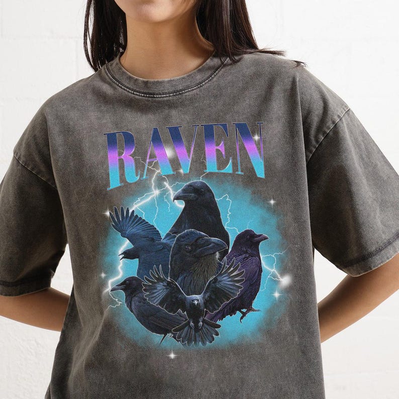 May include: Dark gray t-shirt with a graphic of five black ravens and the word "RAVEN" in a gradient font. The design includes lightning bolts and a blue background, creating a vintage, distressed look.