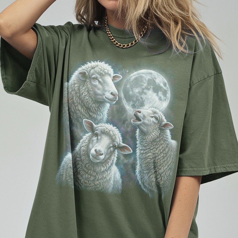 Puede incluir: Camiseta verde oliva con una ilustraci&oacute;n detallada de tres ovejas y una luna llena. Las ovejas est&aacute;n representadas en tonos blancos y grises, con la luna de fondo. La camiseta tiene mangas cortas y un corte holgado.