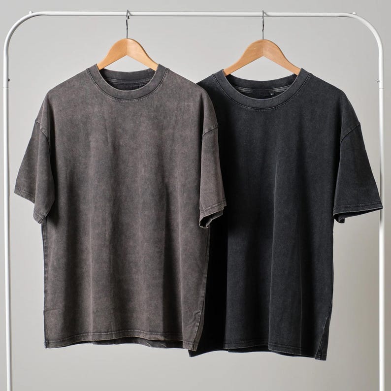 May include: Two oversized, short-sleeved t-shirts in a washed, vintage style. One is a dark charcoal gray, the other is a faded black. Both shirts hang on wooden hangers against a neutral background, showcasing a casual, streetwear aesthetic.