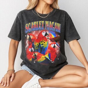 May include: Dark gray t-shirt featuring a vibrant graphic of scarlet macaws with the text "SCARLET MACAW" in a retro font. The design includes red, yellow, blue, and white parrots with lightning bolt accents, creating a vintage aesthetic.