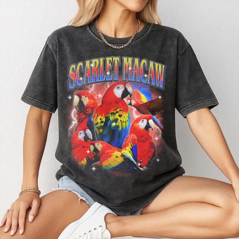 May include: Dark gray t-shirt featuring a vibrant graphic of scarlet macaws with the text "SCARLET MACAW" in a retro font. The design includes red, yellow, blue, and white parrots with lightning bolt accents, creating a vintage aesthetic.