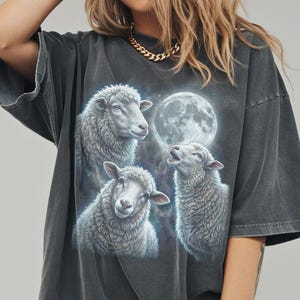 Puede incluir: Camiseta gris oscuro con un gr&aacute;fico de tres ovejas y una luna llena. Las ovejas son blancas con texturas de lana detalladas. La luna es brillante y redonda, sobre un fondo estrellado. La camiseta tiene mangas cortas y un corte holgado.