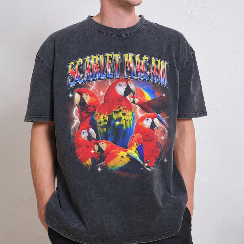 May include: Dark grey t-shirt featuring a vibrant graphic of scarlet macaws with the text "SCARLET MACAW" in a colorful, retro font. The design includes lightning bolts and a cluster of the colorful birds.