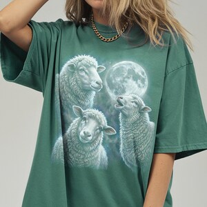 Puede incluir: Camiseta verde con una ilustraci&oacute;n detallada de tres ovejas y una luna llena. Las ovejas est&aacute;n representadas en blanco y gris, con la luna de fondo. La camiseta tiene mangas cortas y un corte holgado.