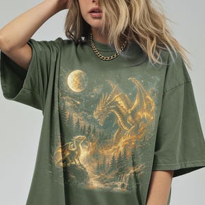 May include: Olive green t-shirt featuring a fantasy scene with two dragons, a full moon, and a forest. The larger dragon is golden and fiery, while the smaller one is light-colored. The design is detailed and artistic.