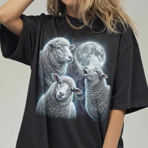 Puede incluir: Camiseta negra con un gr&aacute;fico de tres ovejas y una luna llena. Las ovejas son blancas con texturas de lana detalladas, y la luna es brillante sobre un fondo estrellado. La camiseta es de color oscuro.