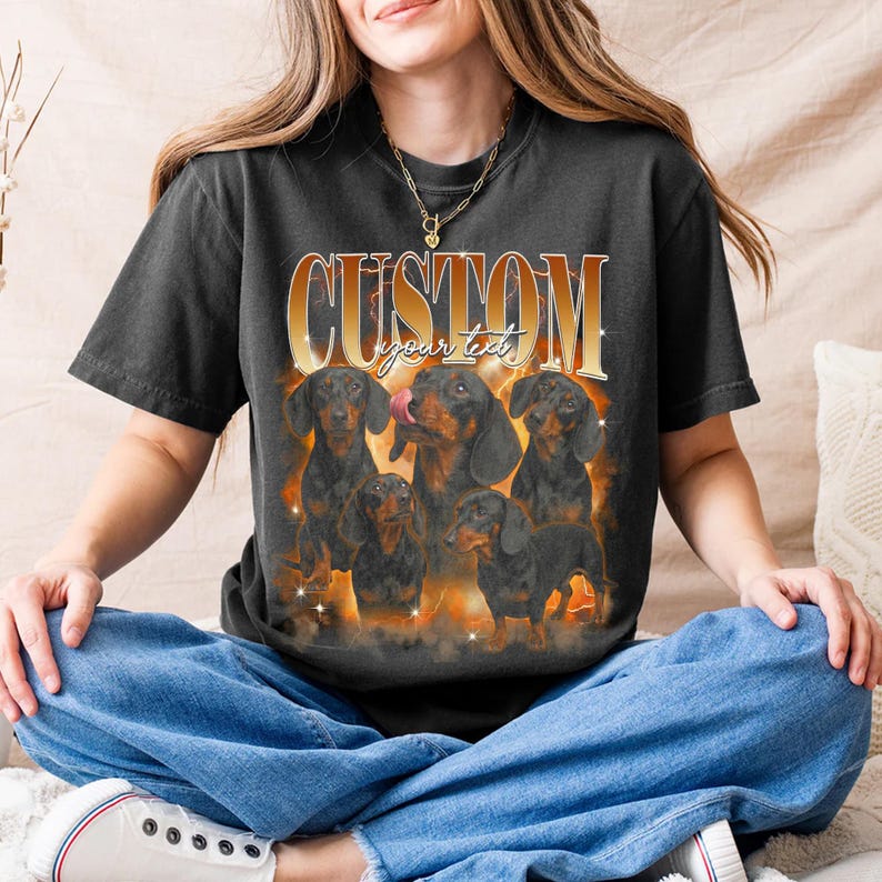 May include: Dark grey t-shirt with a graphic of six dachshunds and a fiery background. The gold text reads "CUSTOM" above "your text". The shirt has short sleeves and is a casual style.