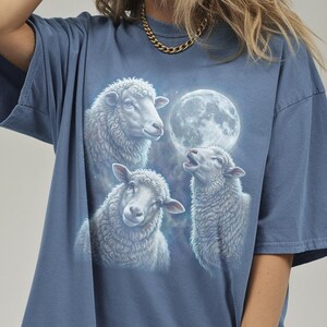 Puede incluir: Camiseta azul con una ilustraci&oacute;n detallada de tres ovejas y una luna llena. Las ovejas est&aacute;n representadas con lana blanca y esponjosa, y la luna es un orbe brillante en el fondo. La camiseta es de un estilo c&oacute;modo e informal.