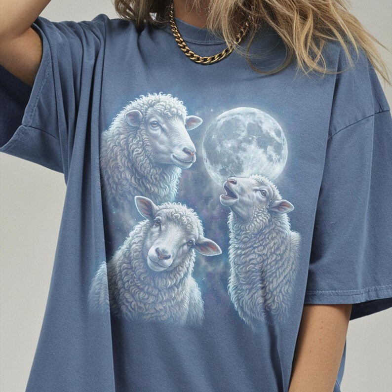 Puede incluir: Camiseta azul con una ilustraci&oacute;n detallada de tres ovejas y una luna llena. Las ovejas est&aacute;n representadas con lana blanca y esponjosa, y la luna es un orbe brillante en el fondo. La camiseta es de un estilo c&oacute;modo e informal.