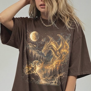 May include: Brown t-shirt featuring a fantasy scene with two dragons, a full moon, and a waterfall. The dragons are gold and the background is a dark night sky with trees. The shirt has short sleeves and a relaxed fit.