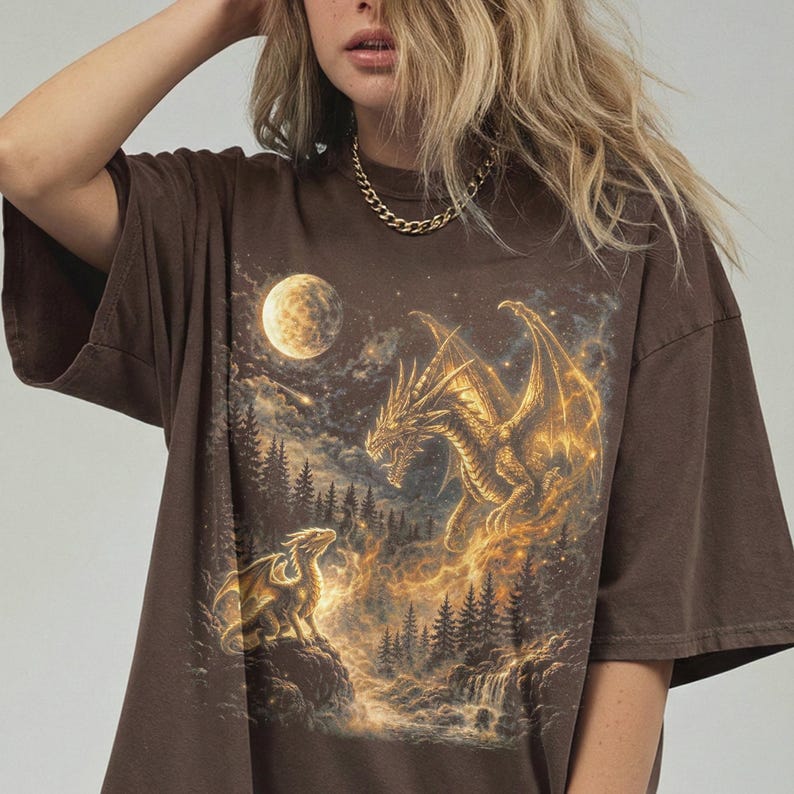 May include: Brown t-shirt featuring a fantasy scene with two dragons, a full moon, and a waterfall. The dragons are gold and the background is a dark night sky with trees. The shirt has short sleeves and a relaxed fit.