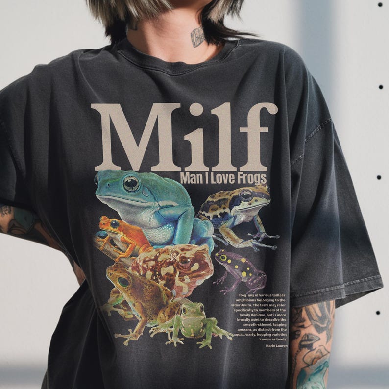 Dark grey acid wash t-shirt featuring the text MILF Man I Love Frogs with vintage scientific illustrations of green tree frogs and spotted toads in a collage style.