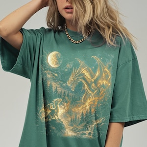 May include: Green t-shirt featuring a fantasy scene with two dragons, a full moon, and a forest. The larger dragon is golden, with the smaller one perched on a rock. The design has a mystical, ethereal quality.