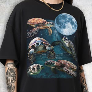 Könnte beinhalten: Schwarzes T-Shirt mit einer farbenfrohen Grafik von fünf Meeresschildkröten und einem großen blauen Mond. Die Schildkröten haben verschiedene Braun-, Grün- und Blautöne. Das Design ist vor einem dunkelblauen, ozeanartigen Hintergrund platziert.