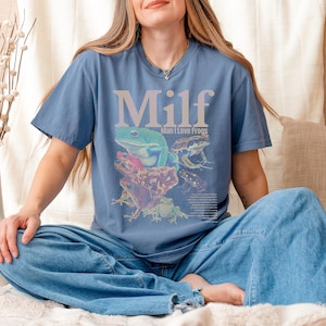 May include: Blue t-shirt with the word "Milf" in large, light purple letters above a graphic of several colorful frogs. The text "Man I Love Frogs" is below the word "Milf". The shirt is made of a soft material.
