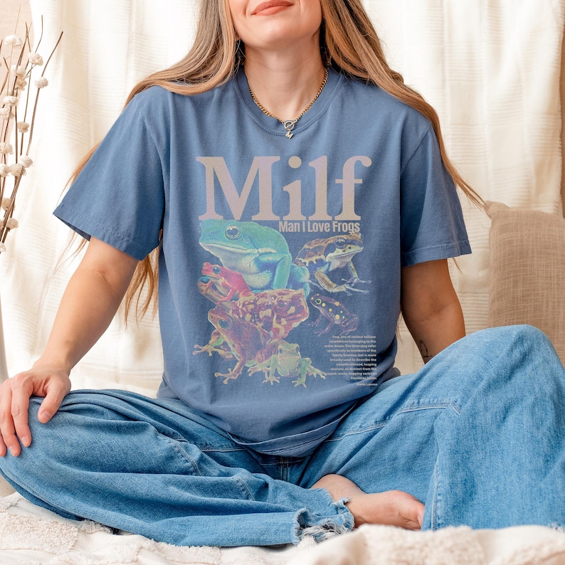 May include: Blue t-shirt with the word "Milf" in large, light purple letters above a graphic of several colorful frogs. The text "Man I Love Frogs" is below the word "Milf". The shirt is made of a soft material.