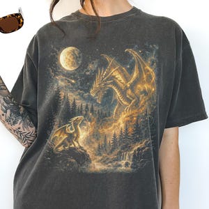 May include: Dark gray t-shirt featuring a fantasy scene with two golden dragons, a full moon, and a forest. The dragons are set against a backdrop of a waterfall and trees, with a glowing, ethereal effect.