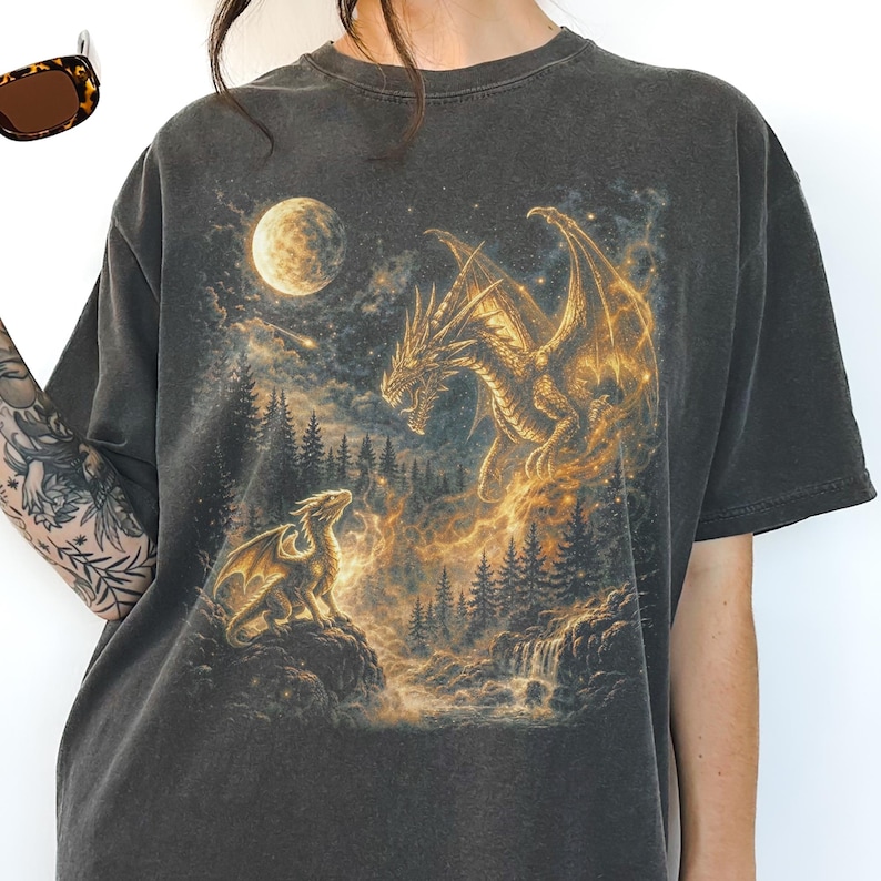 May include: Dark gray t-shirt featuring a fantasy scene with two golden dragons, a full moon, and a forest. The dragons are set against a backdrop of a waterfall and trees, with a glowing, ethereal effect.