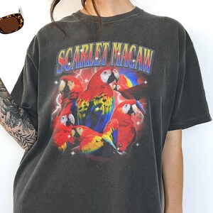 May include: Dark gray t-shirt featuring a vibrant graphic of scarlet macaws. The design includes the text "SCARLET MACAW" in a retro font, with red, yellow, and blue parrots set against a lightning bolt background. A pair of sunglasses is visible in the upper left corner.