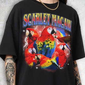May include: Black t-shirt featuring a vibrant graphic of scarlet macaws with red, yellow, and blue plumage. The text "SCARLET MACAW" is arched across the top in a retro font, with a lightning bolt background.