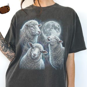 Puede incluir: Camiseta gris oscuro con una ilustraci&oacute;n detallada de tres ovejas y una luna llena. Las ovejas est&aacute;n en tonos grises y blancos, con un aspecto suave y texturizado. La camiseta tiene un aspecto vintage y desgastado.