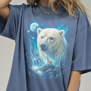 May include: A blue t-shirt with a detailed illustration of a polar bear and cub, a waterfall, and a moonlit sky. The artwork is in white, blue, and green, creating a serene, nature-inspired design. The shirt has short sleeves.