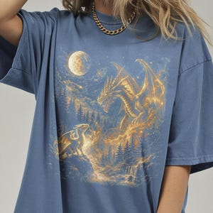 May include: Blue t-shirt featuring a fantasy scene with two dragons, a full moon, and a forest. The dragons are golden and detailed, set against a night sky with a waterfall. The shirt is a comfortable, casual style.