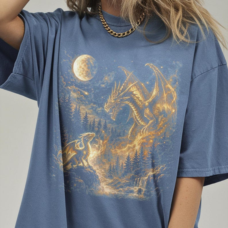 May include: Blue t-shirt featuring a fantasy scene with two dragons, a full moon, and a forest. The dragons are golden and detailed, set against a night sky with a waterfall. The shirt is a comfortable, casual style.