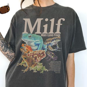 May include: Dark gray t-shirt featuring a colorful frog graphic with the text "Milf Man I Love Frogs". The graphic includes various frog species in shades of green, blue, orange, and brown.