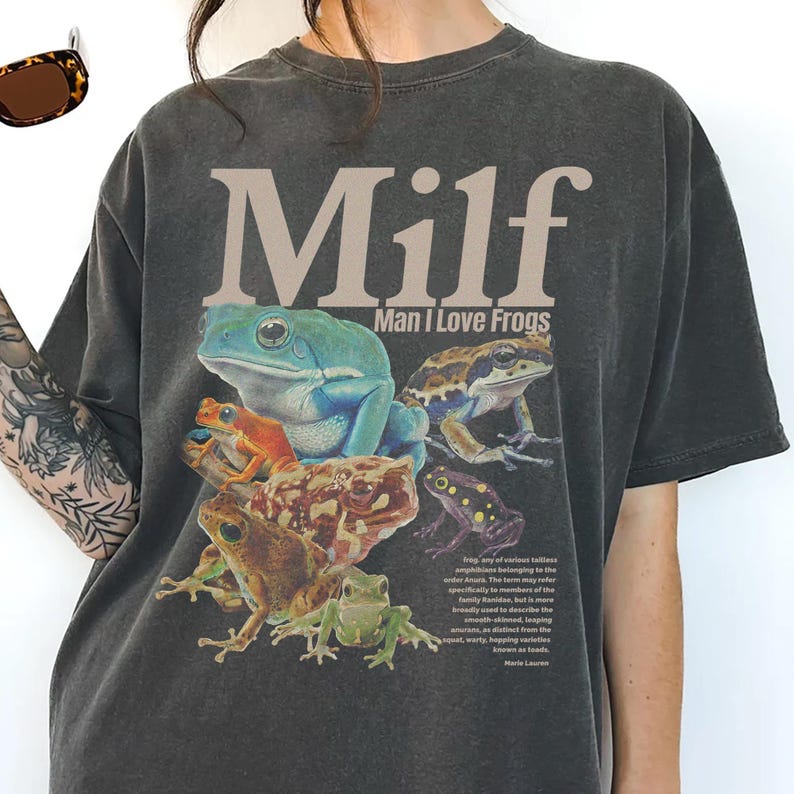 May include: Dark gray t-shirt featuring a colorful frog graphic with the text "Milf Man I Love Frogs". The graphic includes various frog species in shades of green, blue, orange, and brown.