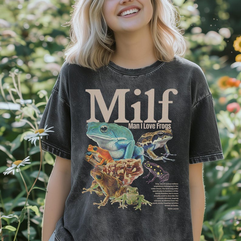 May include: Dark gray t-shirt featuring a graphic of several colorful frogs. The shirt has the word "Milf" in large, bold letters, with "Man I Love Frogs" underneath. The shirt is made of a soft, comfortable material.