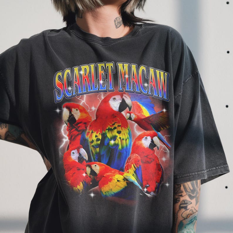 May include: Dark gray t-shirt featuring a vibrant graphic of scarlet macaws. The design includes the text "SCARLET MACAW" in a colorful, retro font, with lightning bolts and stars. The macaws are depicted in red, yellow, blue, and white.