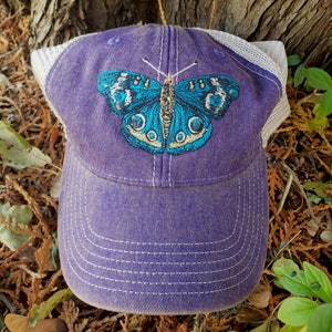 Puede incluir: Gorra de b&eacute;isbol de malla blanca y morada con un dise&ntilde;o de mariposa azul bordada.