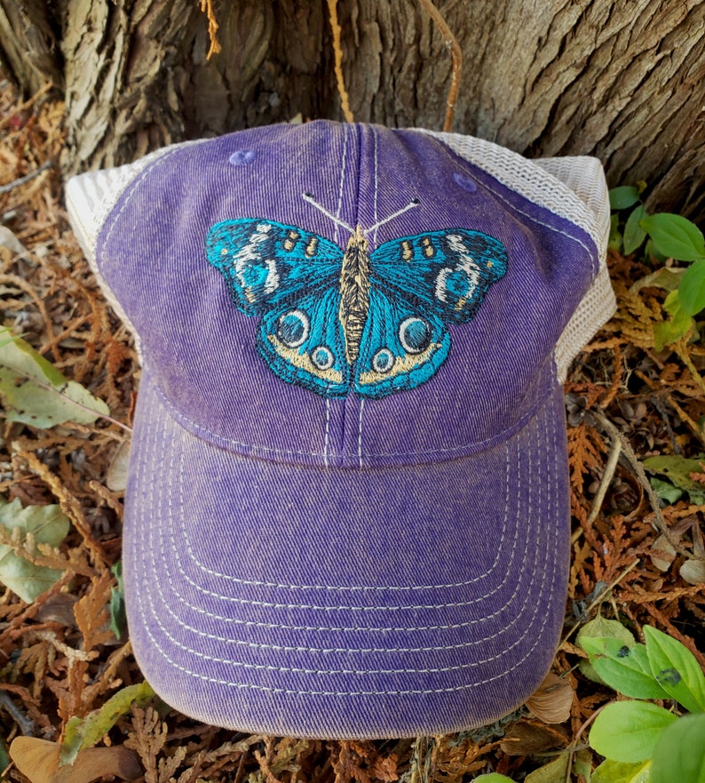 Puede incluir: Gorra de b&eacute;isbol de malla blanca y morada con un dise&ntilde;o de mariposa azul bordada.
