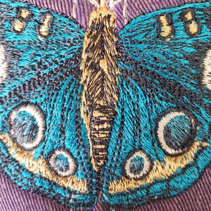 Puede incluir: Primer plano de una mariposa azul bordada en una tela morada. La mariposa tiene detalles en negro y dorado y grandes ojos redondos.