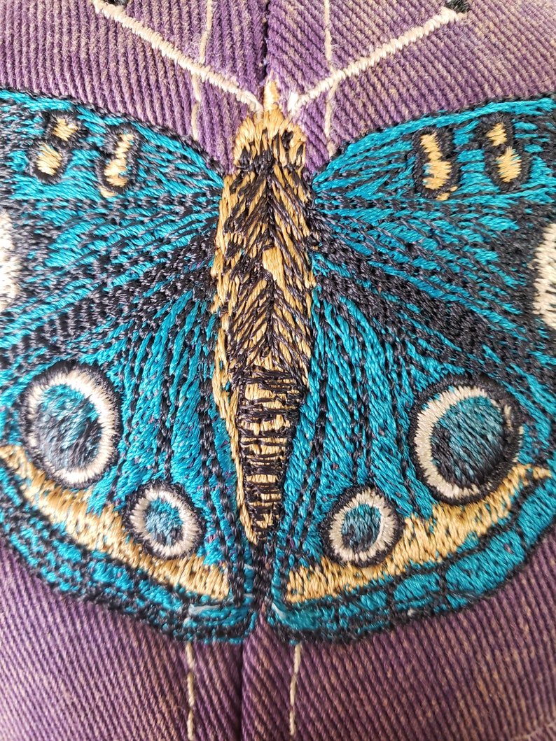 Puede incluir: Primer plano de una mariposa azul bordada en una tela morada. La mariposa tiene detalles en negro y dorado y grandes ojos redondos.