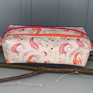 Könnte beinhalten: Eine rechteckige Stofftasche mit einem sich wiederholenden Muster aus Einhörnern und floralen Elementen in Rosa- und Orangetönen. Die Tasche verfügt über einen leuchtend rosa Reißverschluss oben und einen orangefarbenen Reißverschluss vorne.