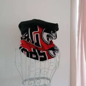Könnte beinhalten: Ein schwarzes und rotes Strickmützen- und Halstuch-Set. Die Mütze ist schwarz, und das Halstuch ist rot mit schwarzen und weißen Buchstaben und einem grafischen Design. Das Set wird auf einer weißen Drahtpuppe präsentiert.