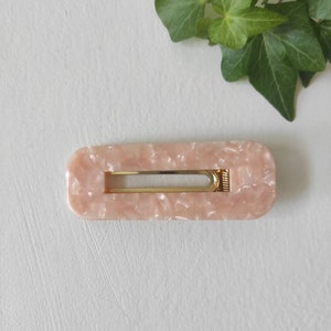May include: A light pink rectangular hair clip with a gold metal bar and a marbled pattern.