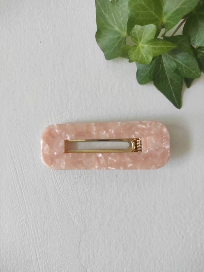 May include: A light pink rectangular hair clip with a gold metal bar and a marbled pattern.