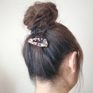 May include: A woman with brown hair in a messy bun, secured with a black scrunchie. A colorful, multi-colored, plastic hair clip is holding a section of hair in place.