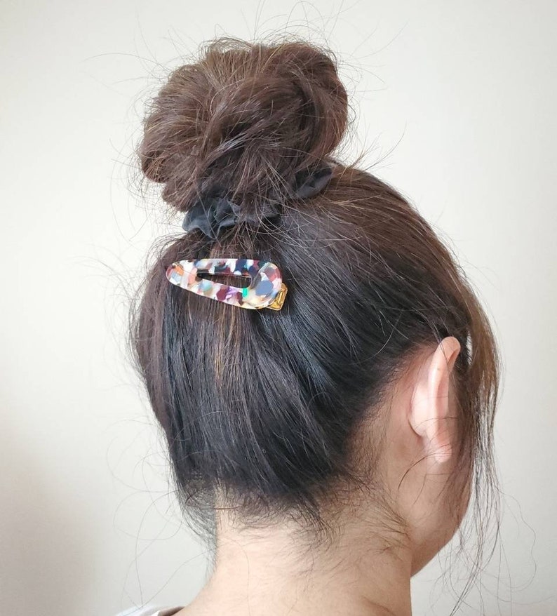 May include: A woman with brown hair in a messy bun, secured with a black scrunchie. A colorful, multi-colored, plastic hair clip is holding a section of hair in place.