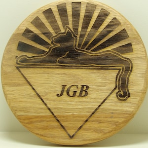 Könnte beinhalten: Eine runde Holzplakette mit einer schwarzen Katzensilhouette in einem Dreieck. Die Katze liegt mit ihrem Schwanz um den Körper gewickelt. Die Plakette hat ein Sonnenstrahlen-Design hinter der Katze und die Initialen "JGB" in der Mitte des Dreiecks.