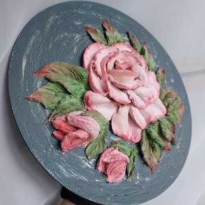 May include: A round, grey canvas with a 3D sculpted floral design. The flowers are pink and white with green leaves.