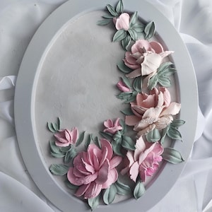 May include: Oval-shaped, gray decorative wall art featuring a floral relief design. The relief includes pink and light pink flowers with green leaves. The flowers are arranged in a curved pattern along the edge of the oval.