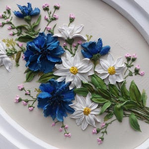 Puede incluir: Primer plano de un marco ovalado blanco con una pintura floral en 3D. La pintura presenta flores azules y blancas con hojas verdes y capullos rosados.