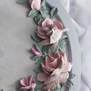 May include: A decorative, round, gray tray with a textured surface. The tray is adorned with a floral design featuring pink and off-white roses, and green leaves. The flowers are sculpted, creating a 3D effect.