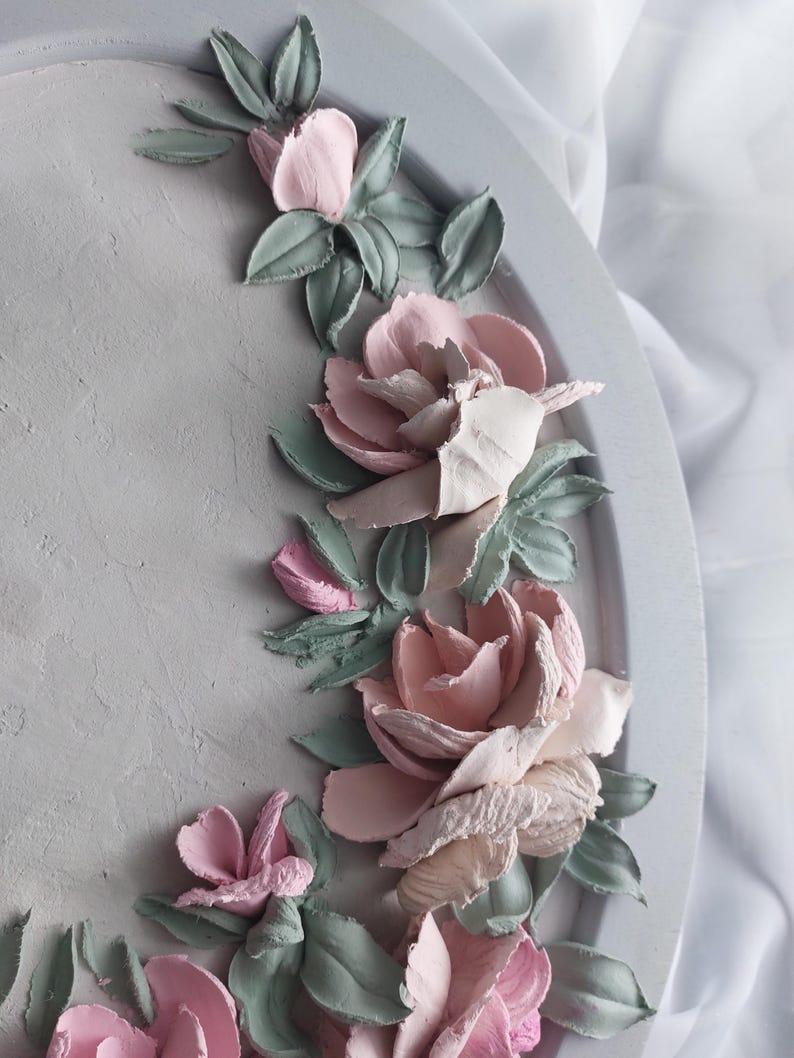 May include: A decorative, round, gray tray with a textured surface. The tray is adorned with a floral design featuring pink and off-white roses, and green leaves. The flowers are sculpted, creating a 3D effect.
